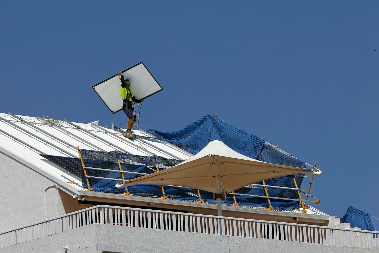 An installer in hi-vis and a harness carries a solar panel across a white metal roof behind safety rails